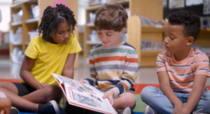 3 children sitting on the floor of a library reading a book together.