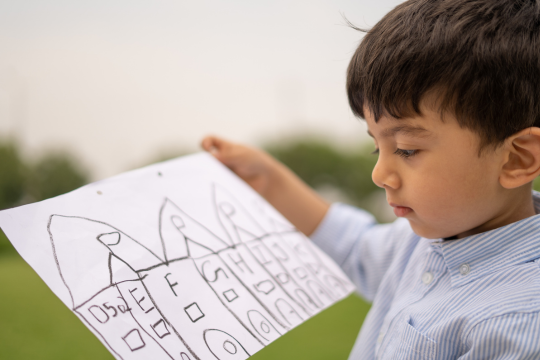 A young boy wearing a button up shirt holds a drawing of a row of houses.