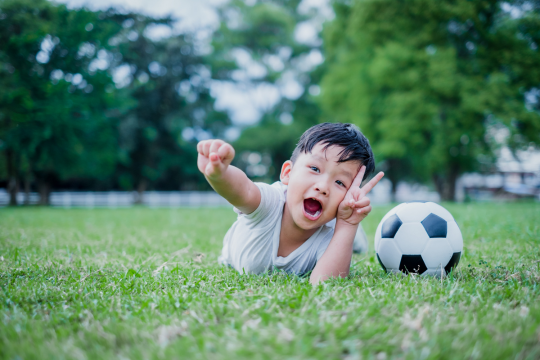 A young boy lays in the grass making the peace sign with his hand. He has a soccer ball.