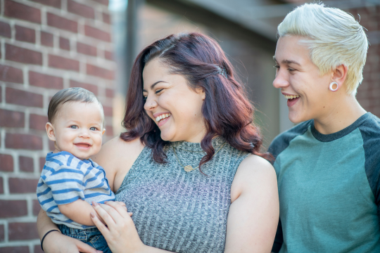One woman with short auburn hair holds a baby, while another woman (her partner) with short blonde hair looks on and smiles.